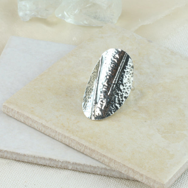 Silver ring with a large oval shape that wraps around the finger. It features two folds and a hammered texture. The oval has an oxidised darker finish. The round silver ring band is adjustable in size.