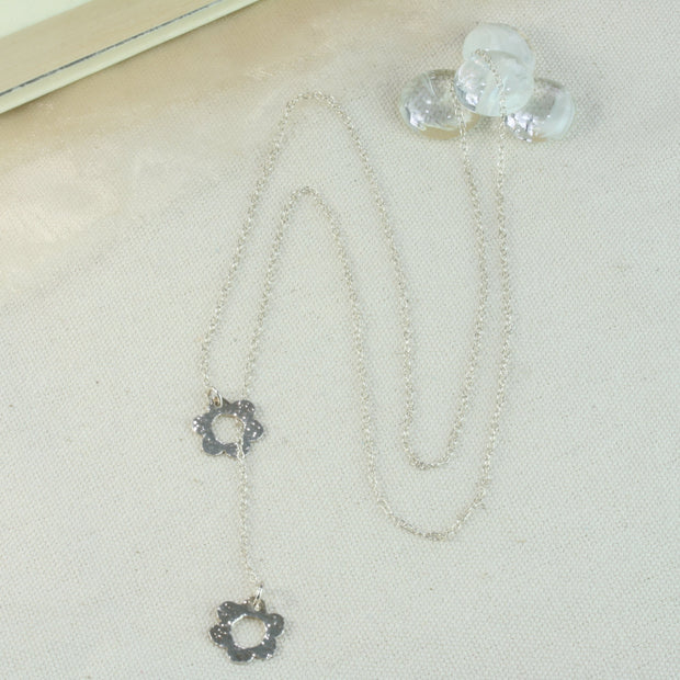 Silver y necklace featuring two flowers. The chain is looped through one flower, the other flower dangles off the other end of the chain. Both flowers have a hammered shiny finish. There are two variants, one with two flowers of the same size and the other featuring a larger flower at the top and a smaller one at the bottom.