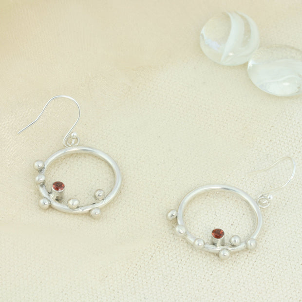Silver hoop hook earrings featuring 6 silver balls and a Garnet gemstone. The gemstone is 3mm in diameter and is set in a tube setting at the bottom and slightly to one side on the inside of the hoop. These are handmade earrings, made form eco-silver.
