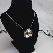Silver pendant necklace featuring a circle that has been cut in half. Both sides have been domed into half a cup shape and soldered together with the dome on different sides. One side has a striped texture the other a round hammered texture. It has a mirror finish. The snake chain necklace is 45 cm / 18" long.
