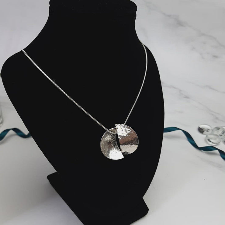 Silver pendant necklace featuring a circle that has been cut in half. Both sides have been domed into half a cup shape and soldered together with the dome on different sides. One side has a striped texture the other a round hammered texture. It has a mirror finish. The snake chain necklace is 45 cm / 18" long.