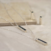 Eco silver pendant necklace featuring two bars. Both have a square hammered texture and shiny mirror finish. They are attached to two chain with different lengths letting one bar sitting above the other. Seen here with the eco silver bar stud earrings in the background.