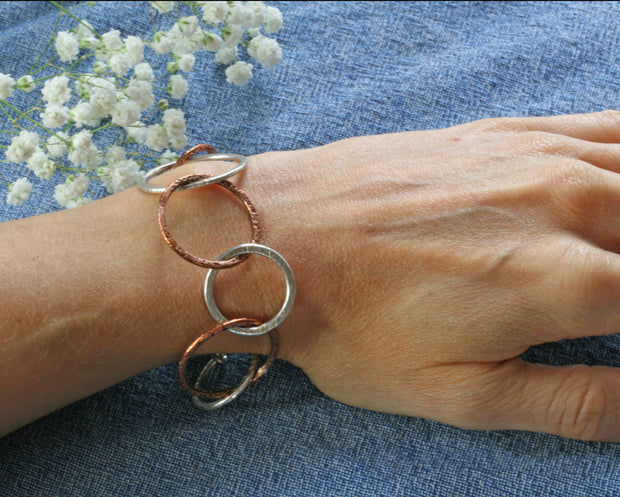 The matching silver bracelet featuring silver hoops in various sizes and various hammered textures. Four of the nine hoops are made from copper and have a hammered texture as well. The bracelet has a shiny finish.