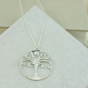 A silver round tree of life pendant necklace. A circle featuring a sawn out unique Tree of Life design. It measures 3 cm in diameter and has round curled branches with the top two branches coming together in a heart shape. It has a shiny hammered texture and the necklace is available in 45 cm and 76 cm lengths.