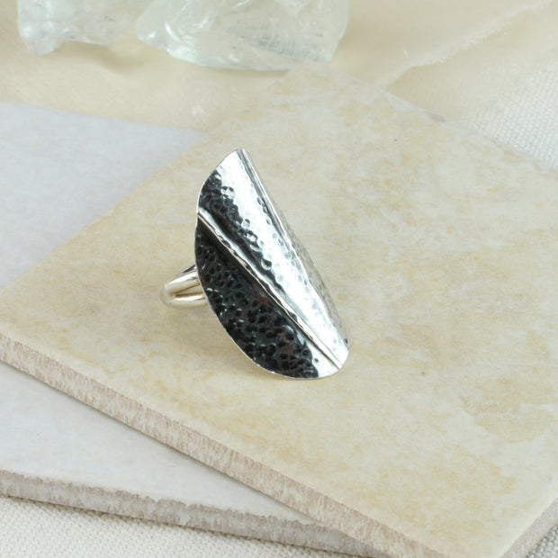 Silver ring with a large oval shape that wraps around the finger. It features two folds and a hammered texture. The oval has an oxidised darker finish. The round silver ring band is adjustable in size.