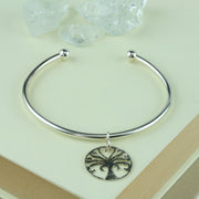 Silver bangle with a silver Tree of Life charm. A silver ball on each end of the bangle keeps the charm in place. The charm has a textured finish on one side and a mirror finish on the other.