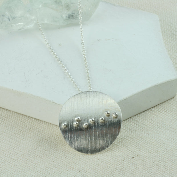 Eco silver circle pendant with a striped texture. 8 silver balls are placed in a slight diagonal line at random spots from left to right. The circle is slightly arched and has a mirror finish.
