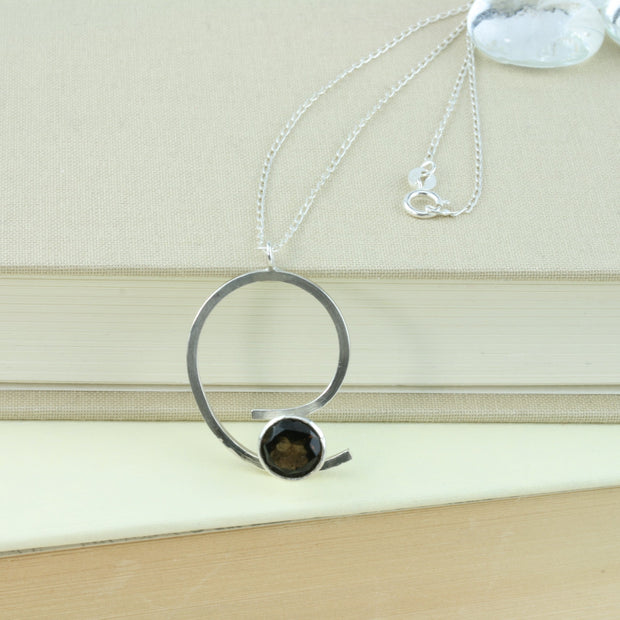 Eco silver swirl pendant with a collet setting held by either end of the swirl. The collet setting features a Smoky Quartz gemstone. The collet setting is open at the back allowing light to fall through and brighten up the gemstone. This has a smoky brown grey colour depending on how it catches the light.