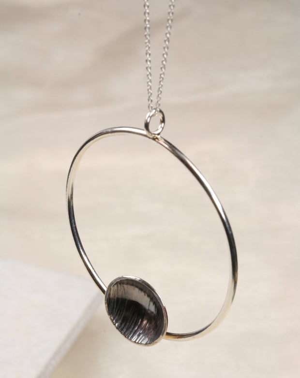 Eco silver hoop pendant necklace with dish. The hoop has a shiny mirror finish and features a silver domed dish on the inside bottom. The dish has a hammered texture and the inside is oxidised and polished to show a darker finish in contrast to the hoop.