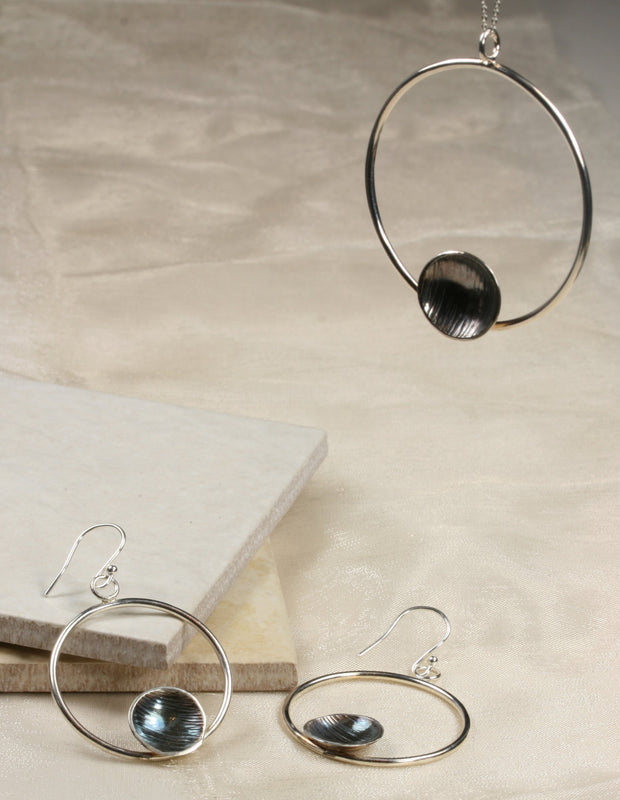 Eco silver hoop earrings with a dish on the inside at the bottom of the hoop. The hoop it attached to a hook earrings at the top. The hoops have a shiny miror finish and the cups have a striped texture with a darker oxidies finsh. The cups are polished to a shine as well. The backs of the cups have a mirror finish.