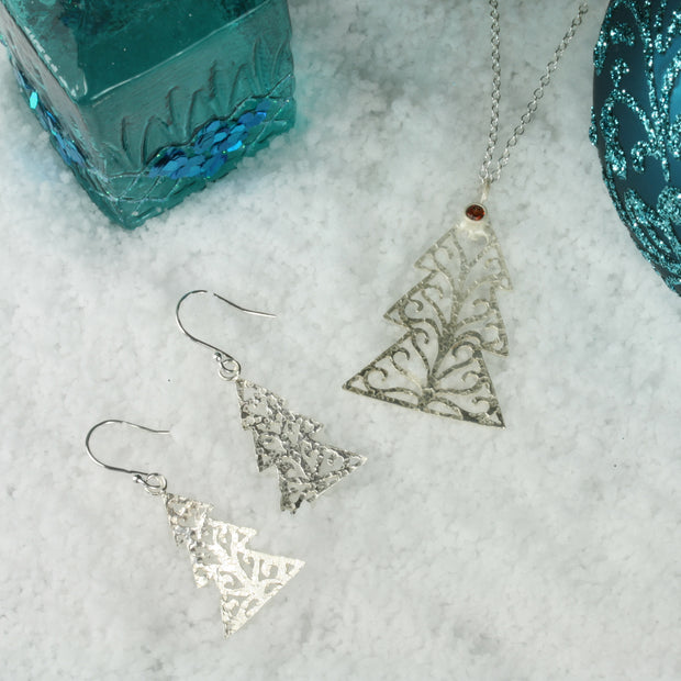 Silver hook earrings in the shape of a Christmas three with three layers. The trees feature a hand drawn pattern which is hand sawn with swirls from the centre to the sides. They have a hammered texture which has been polished to a shiny finish, giving them a shimmer effect.  Seen here with the matching pendant necklace featuring a Garnet gemstone at the top of the tree.