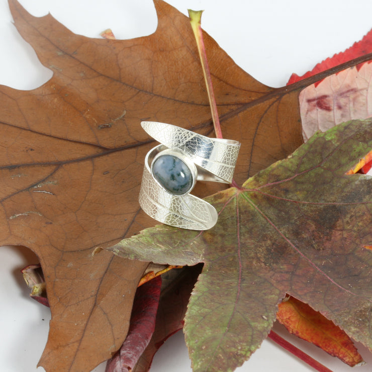 Eco silver leaf adjustable ring. A ring band in a long leaf shape with a real leaf texture. Both ends are sitting parallel to each other to hold the Moss Agate gemstone between them. The gemsotne is attached to the band on one side to give the ring its adjustable feature, by pushing or pulling on the ring slightly.