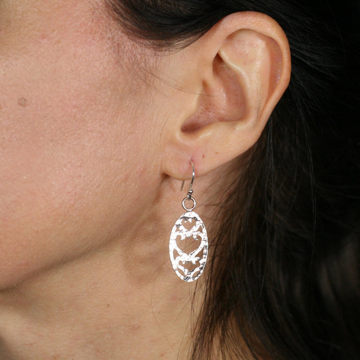 Silver hook earrings featuring an oval shape with three heart shapes cut out of the centre. The middle heart is larger than the two hearts above and below it. The hook has a little silver ball on the front. The ovals have a hammered shiny finish.