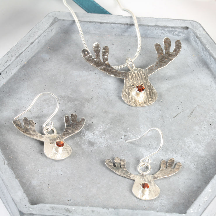 Silver pendant necklace featuring a sawn out Rudolph head. The face has a striped texture and shiny silver finish. The Antlers have a round hammered texture and are aged to give them a darker look, they have been polished to a shine. The red nose is a Garnet gemstone set in a tube setting. Matching hook earrings and ring are available as well.