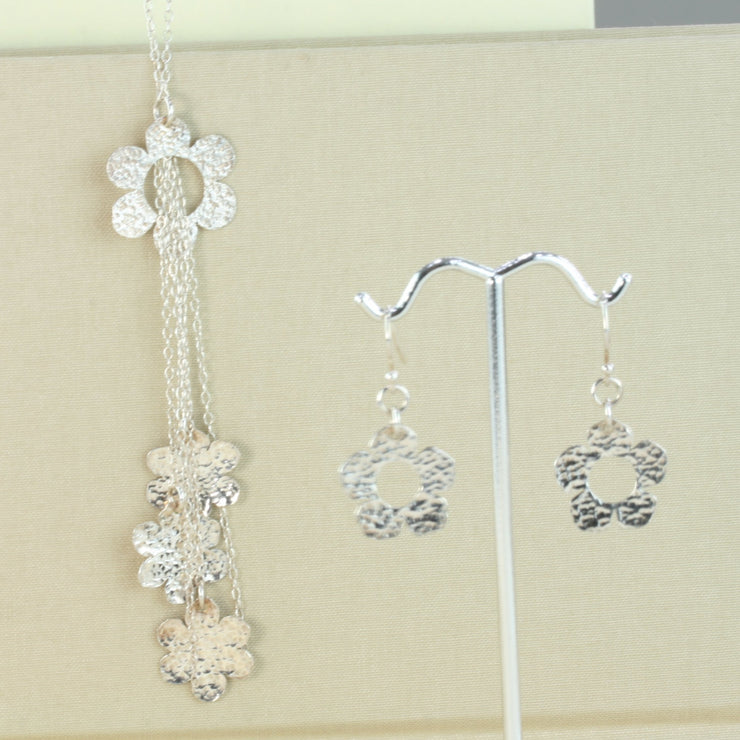 Silver necklace featuring one open flower with five strands of chain looped through the centre. Three of the chains of different lengths feature a smaller flower. All flowers have a hammered texture and shiny finish. Seen here with the silver flower hook earrings.