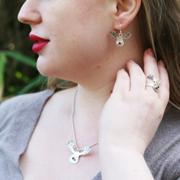 Silver pendant necklace featuring a sawn out Rudolph head. The face has a striped texture and shiny silver finish. The Antlers have a round hammered texture and are aged to give them a darker look, they have been polished to a shine. The red nose is a Garnet gemstone set in a tube setting. Matching hook earrings and ring are available as well.