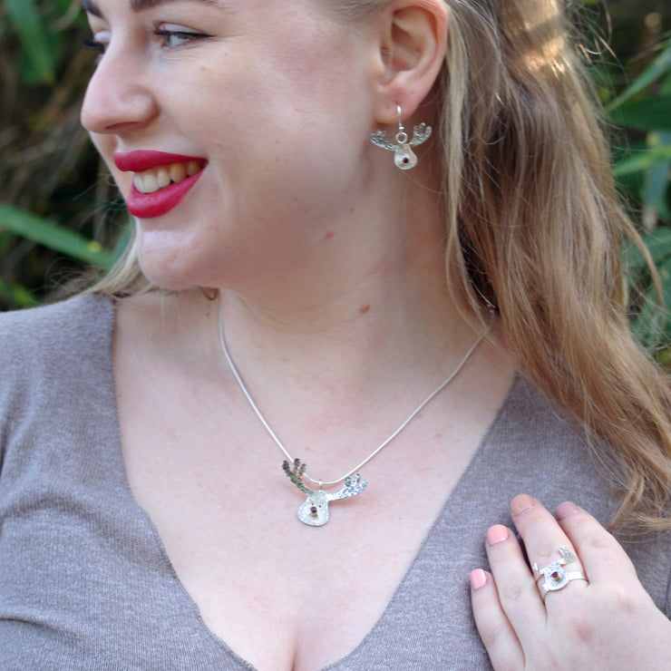 Silver pendant necklace featuring a sawn out Rudolph head. The face has a striped texture and shiny silver finish. The Antlers have a round hammered texture and are aged to give them a darker look, they have been polished to a shine. The red nose is a Garnet gemstone set in a tube setting. Matching hook earrings and ring are available as well.