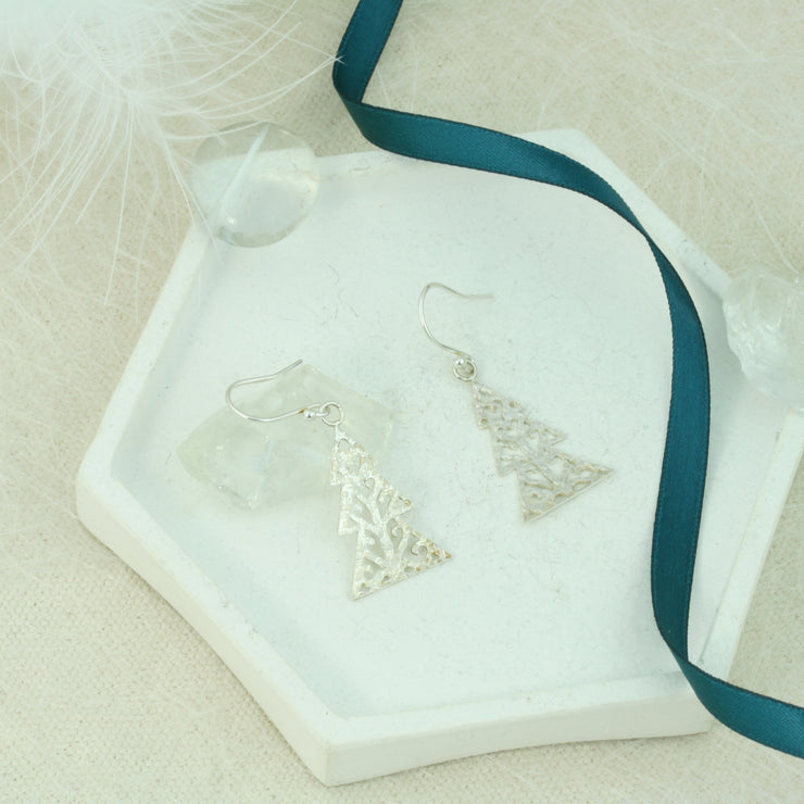 Silver hook earrings in the shape of a Christmas three with three layers. The trees feature a hand drawn pattern which is hand sawn with swirls from the centre to the sides. They have a hammered texture which has been polished to a shiny finish, giving them a shimmer effect. 