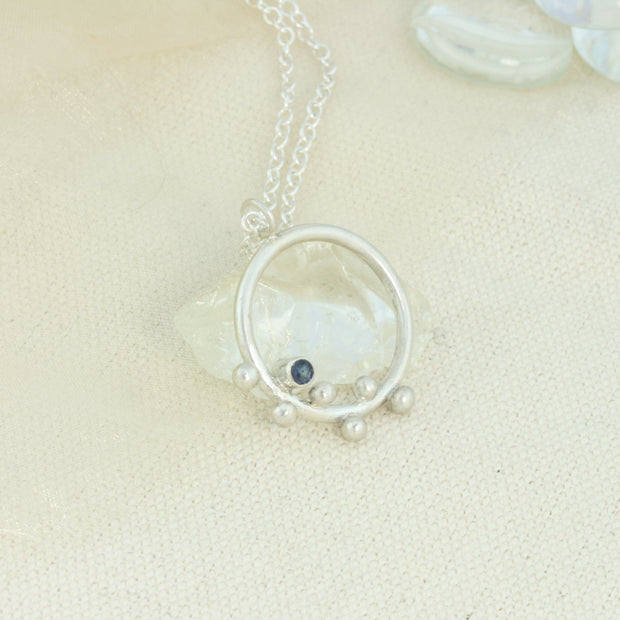 Silver hoop pendant featuring 6 silver balls and a Sapphire gemstone. The gemstone is 3mm in diameter and is set in a tube setting at the bottom left side on the inside of the hoop. This is a handmade pendant, made form eco-silver. The trace chain necklace can be fastened with its lobster clasp at either 40cm, 45cm or 50cm.