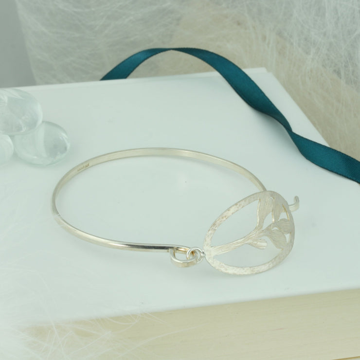 Bangle bracelet with both ends fastened by the feature Mistletoe shape. The Mistletoe is framed byan oval shape and both have a hammered texture, the Mistkletoe has a striped finsh, the oval frame a round hammered texture. The bangle itself has a mirror finish. The bangle fastens by looping one end trough the jump ring on the side of the oval frame.