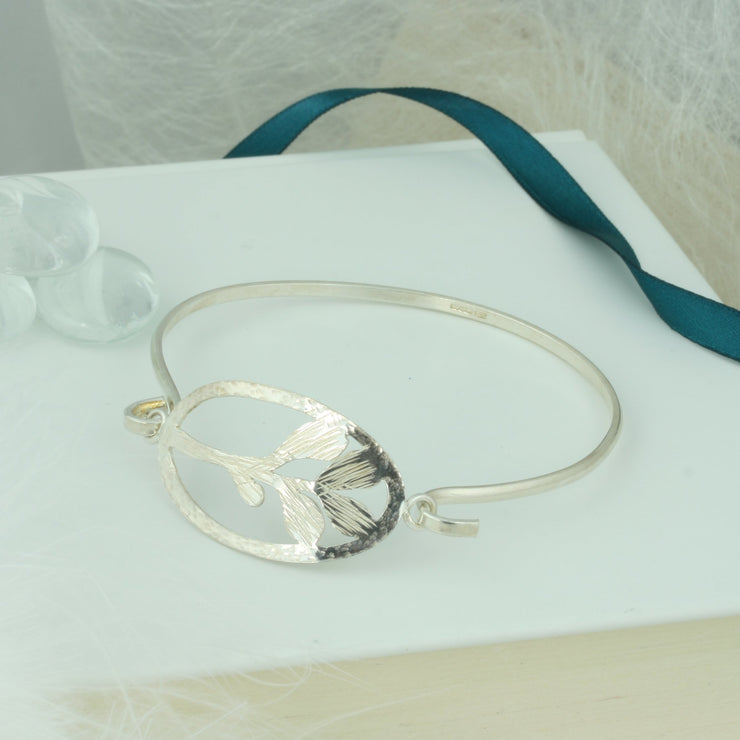 Bangle bracelet with both ends fastened by the feature Mistletoe shape. The Mistletoe is framed byan oval shape and both have a hammered texture, the Mistkletoe has a striped finsh, the oval frame a round hammered texture. The bangle itself has a mirror finish. The bangle fastens by looping one end trough the jump ring on the side of the oval frame.