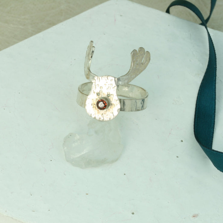 Silver adjustable ring featuring a sawn out Rudolph head. The face has a striped texture and shiny silver finish. The Antlers have a round hammered texture and are aged to give them a darker look, they have been polished to a shine. The red nose is a Garnet gemstone set in a tube setting. The ring band has a striped hammered texture. The ring is adjustable by pinching or pulling the ring band slightly. Matching hook earrings and pendant necklace are available as well.