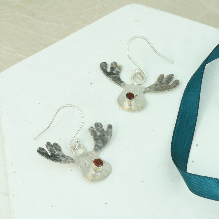 Silver hook earrings featuring a sawn out Rudolph head. The face has a striped texture and shiny silver finish. The Antlers have a round hammered texture and are aged to give them a darker look, they have been polished to a shine. The red nose is a Garnet gemstone set in a tube setting. A matching pendant necklace and ring are available as well. 