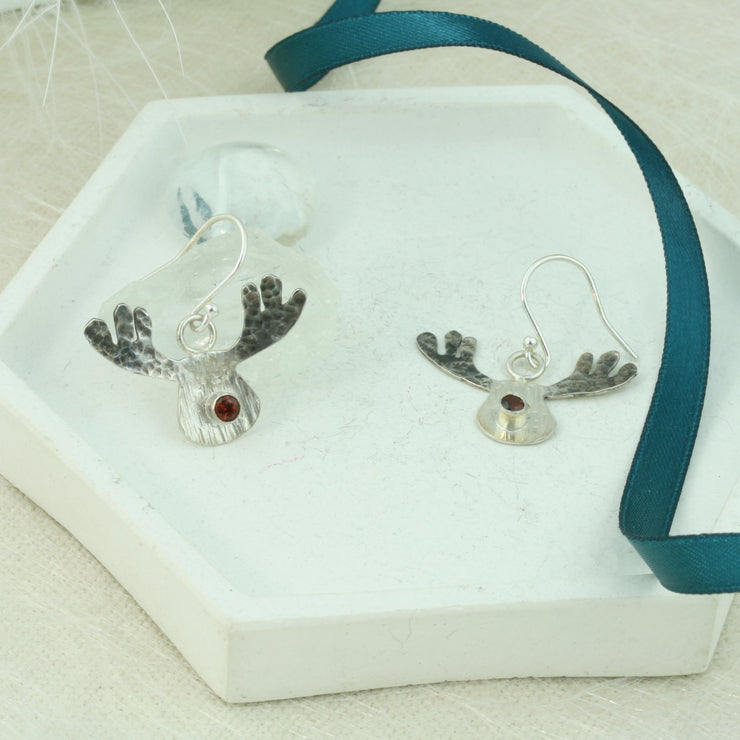 Silver hook earrings featuring a sawn out Rudolph head. The face has a striped texture and shiny silver finish. The Antlers have a round hammered texture and are aged to give them a darker look, they have been polished to a shine. The red nose is a Garnet gemstone set in a tube setting. A matching pendant necklace and ring are available as well. 