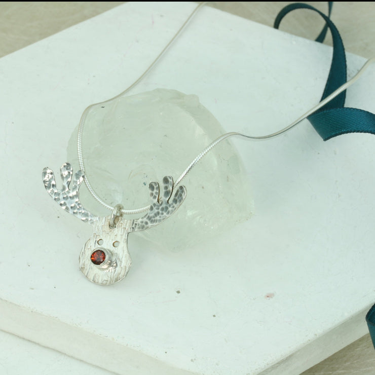 Silver pendant necklace featuring a sawn out Rudolph head. The face has a striped texture and shiny silver finish. The Antlers have a round hammered texture and are aged to give them a darker look, they have been polished to a shine. The red nose is a Garnet gemstone set in a tube setting. Matching hook earrings and ring are available as well.