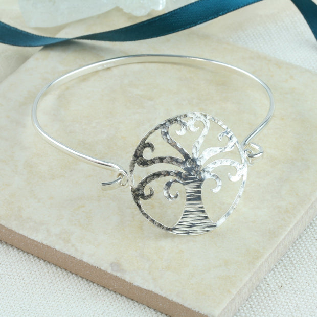 Silver bangle bracelet featuring a TRee of Life at the front. The wire of the bangle is fastened on one side of the Tree of Life. The other side can be looped through the jump ring attached to the Tree of Life to open and close the bangle bracelet. It is made from eco silver and has a shiny finish. The Tree of Life has a hammered texture.