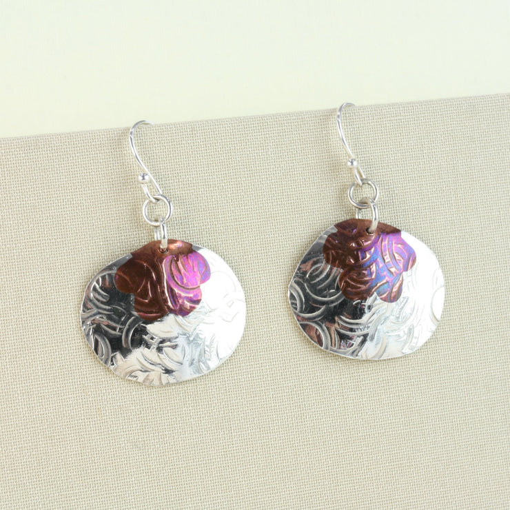 Silver earrings in a large oval shape. Attached to the hooks horizontally, they have a flower shape sawn out of the top which has been filled with a copper inlay. The earring is slightly domed and has a round hammered texture. The copper has a dark brow or purple look depending on how the light hits. The earrings have a shiny mirror finish.