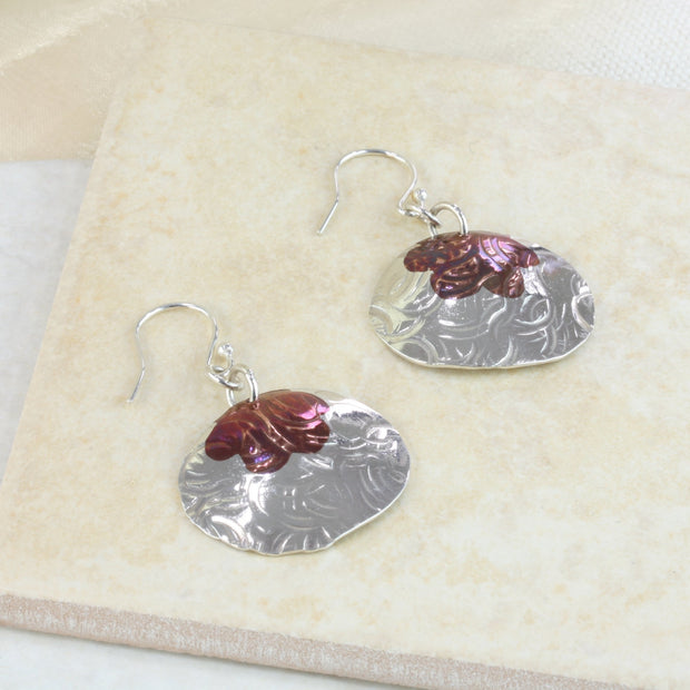 Silver earrings in a large oval shape. Attached to the hooks horizontally, they have a flower shape sawn out of the top which has been filled with a copper inlay. The earring is slightly domed and has a round hammered texture. The copper has a dark brow or purple look depending on how the light hits. The earrings have a shiny mirror finish.