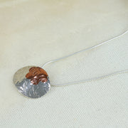 Silver pendant necklace in a large oval shape. The chain is looped through the pendant horizontally, it has a flower shape sawn out of the top which has been filled with a copper inlay. The pendant is slightly domed and has a round hammered texture. The copper has a brow or purple look depending on how the light hits. The pendant has a shiny mirror finish.