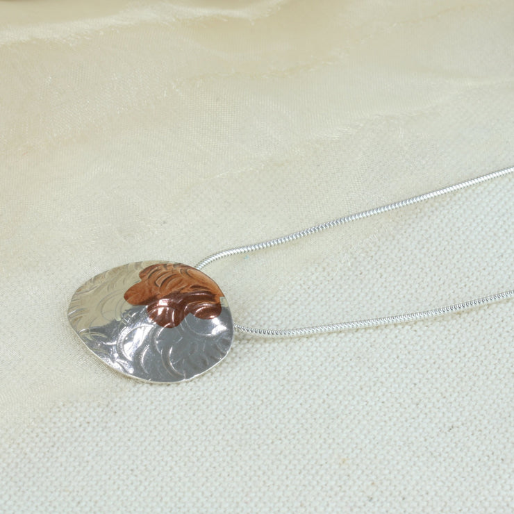 Silver pendant necklace in a large oval shape. The chain is looped through the pendant horizontally, it has a flower shape sawn out of the top which has been filled with a copper inlay. The pendant is slightly domed and has a round hammered texture. The copper has a brow or purple look depending on how the light hits. The pendant has a shiny mirror finish.