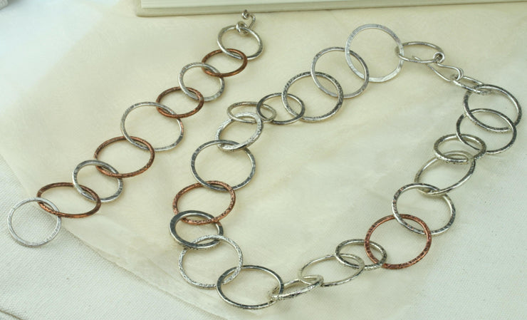 Silver statement necklace featuring silver hoops in various sizes and various hammered textures. Two hoops near the middle of the necklace are made from copper and have a hammered texture as well. The necklace has a shiny finish. Seen here with the matching bracelet.