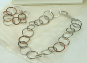 Silver statement necklace featuring silver hoops in various sizes and various hammered textures. Two hoops near the middle of the necklace are made from copper and have a hammered texture as well. The necklace has a shiny finish. Seen here with the matching bracelet.