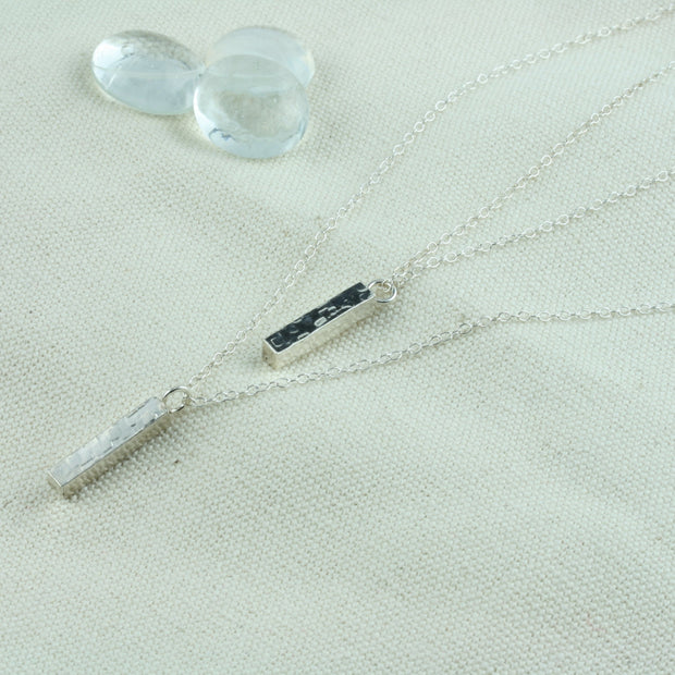 Eco silver pendant necklace featuring two bars. Both have a square hammered texture and shiny mirror finish. They are attached to two chain with different lengths letting one bar sitting above the other.