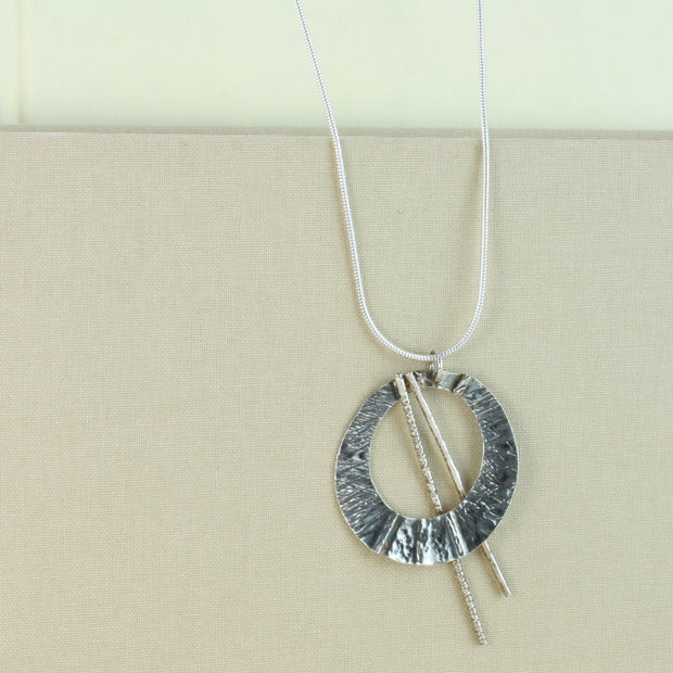Silver open circle pendant with a hammered texture and folds from top to bottom. It has a darker oxidised finish and is open to let two bars through. They have a hammered texture and a shiny silver finish. The necklace is made from snake chain.
