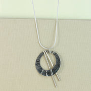 Silver open circle pendant with a hammered texture and folds from top to bottom. It has a darker oxidised finish and is open to let two bars through. They have a hammered texture and a shiny silver finish. The necklace is made from snake chain.