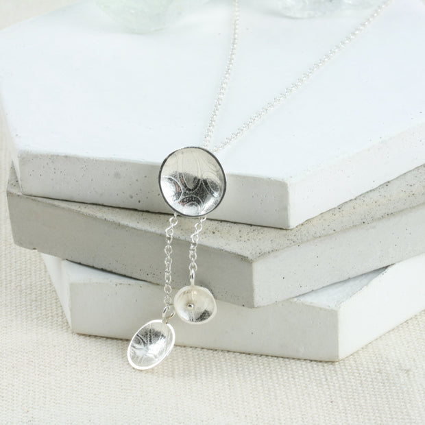 Silver cups drop necklace; a silver necklace featuring a drop pendant with a larger domed cup at the top and two smaller cups attached by chain for a drop effect. One cup is smaller than the other and features a gold ball. It has a pebble texture.