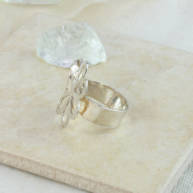 Silver wider ring band that is open on one end and holding the flower in place on the other end. The flower has rounded leaves and the flower and band have a round hammered texture and mirror finish.
