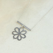 Silver silhouette of a round leaved flower. The flower has a hammered texture and shiny finish. Dangling off a round bar. The chain is looped through the bar and fastens with a lobster clasp.