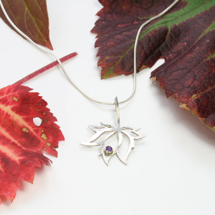 A silver pendant necklace featuring the silhouette of a flower with 3 petals and two leaves on both sides. The middle petal holds an Amethyst gemstone in a tube setting in place, at the tip inside the flower. The necklace is a snake chain with a lobster clasp.