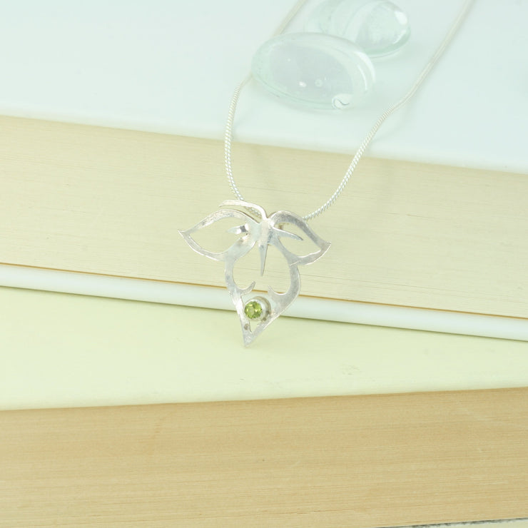 Silver pendant featuring a flower silhouette with a flower bud in the middle and a leaf on either side. The bottom of the flower bud features a peridot gemstone set in a tube setting. It has a mirror finish. The necklace is a snake chain with a lobster clasp fastening.