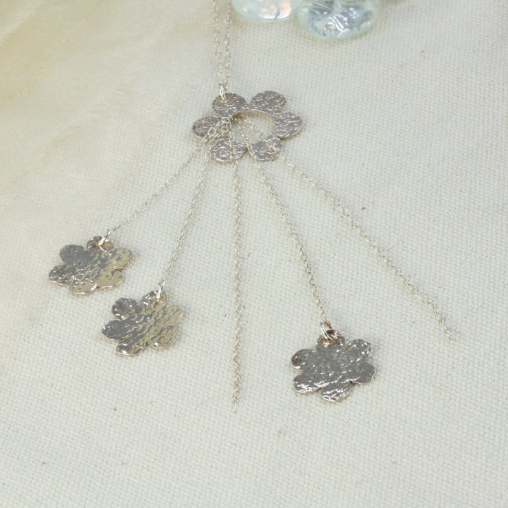 Silver necklace featuring one open flower with five strands of chain looped through the centre. Three of the chains of different lengths feature a smaller flower. All flowers have a hammered texture and shiny finish.