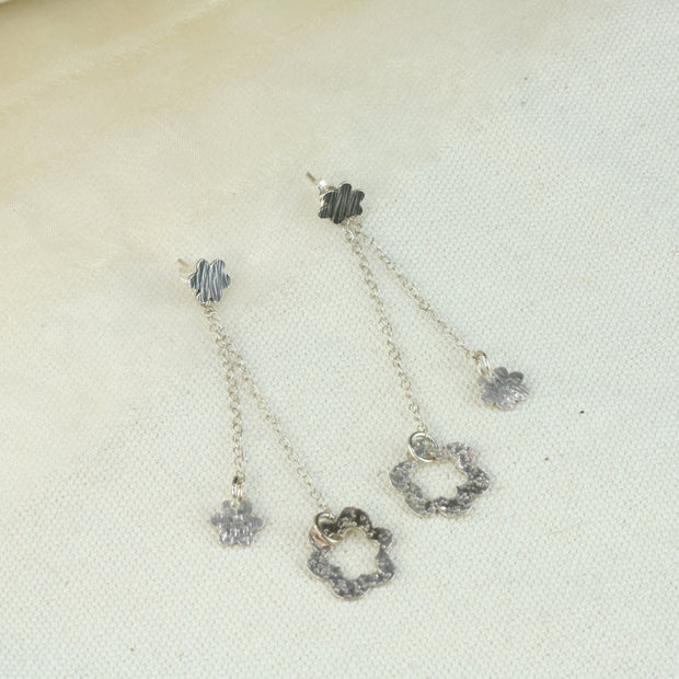 Small silver flower stdus with a striped hammered texture and a darker oxidised finish. A drop part is attached by looping the stud through a jump ring. The drop features a smaller flower and a larger open flower. Both are attached to trace chain with the larger flower a bit lower than the smaller one. Two looks in one.