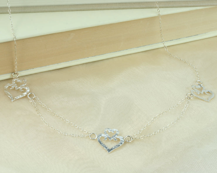 Silver statement necklace. Featuring three hearts shapes with a smaller heart on top of a larger heart as a crown. The heart in the middle is connected to the two hearts on either side with two chains to add a dramatic effect. The two outside hearts are attached to a single trace chain to make up the rest of the necklace. It has a lobtser clasp on one end and a necklace extender on the other side.