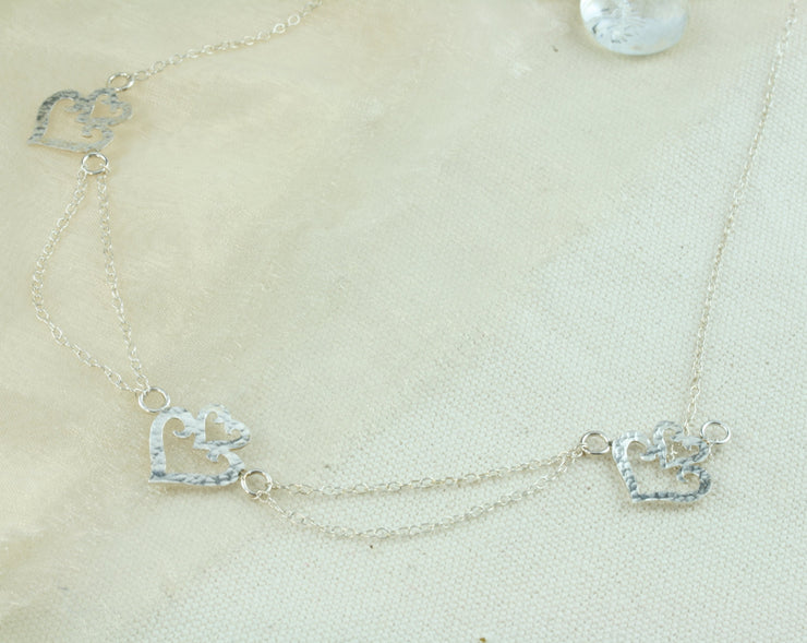 Silver statement necklace. Featuring three hearts shapes with a smaller heart on top of a larger heart as a crown. The heart in the middle is connected to the two hearts on either side with two chains to add a dramatic effect. The two outside hearts are attached to a single trace chain to make up the rest of the necklace. It has a lobtser clasp on one end and a necklace extender on the other side.