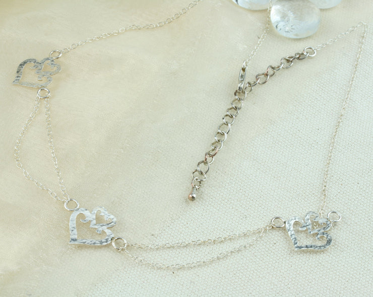 Silver statement necklace. Featuring three hearts shapes with a smaller heart on top of a larger heart as a crown. The heart in the middle is connected to the two hearts on either side with two chains to add a dramatic effect. The two outside hearts are attached to a single trace chain to make up the rest of the necklace. It has a lobtser clasp on one end and a necklace extender on the other side.