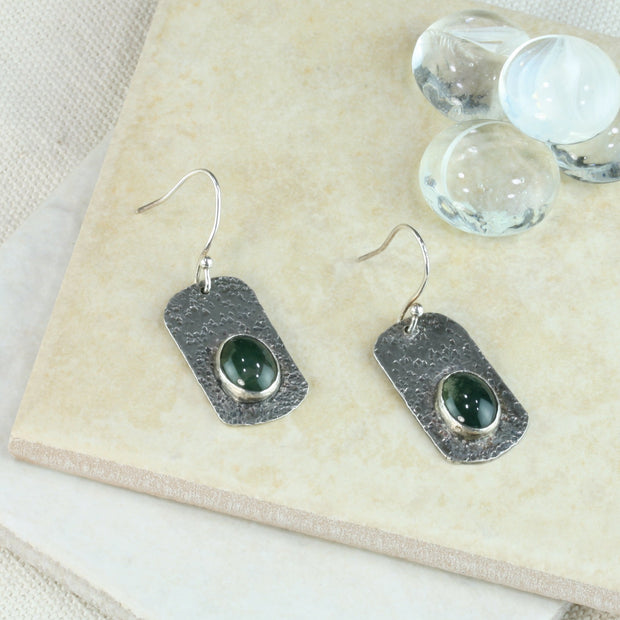 Silver hook earrings featuring a rectangle shape with a round hammered texture. The silver has been oxidised to give it a darker finish. The Moss Agate gemstones are oval and the back behind them has been cut out to let the light shine through , perfectly showing the details of the gemstones.